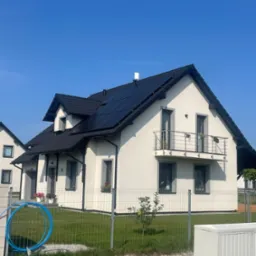 Nowoczesny dom jednorodzinny z balkonem i panelami słonecznymi na dachu, widoczny zza ogrodzenia panelowego, na tle błękitnego nieba.