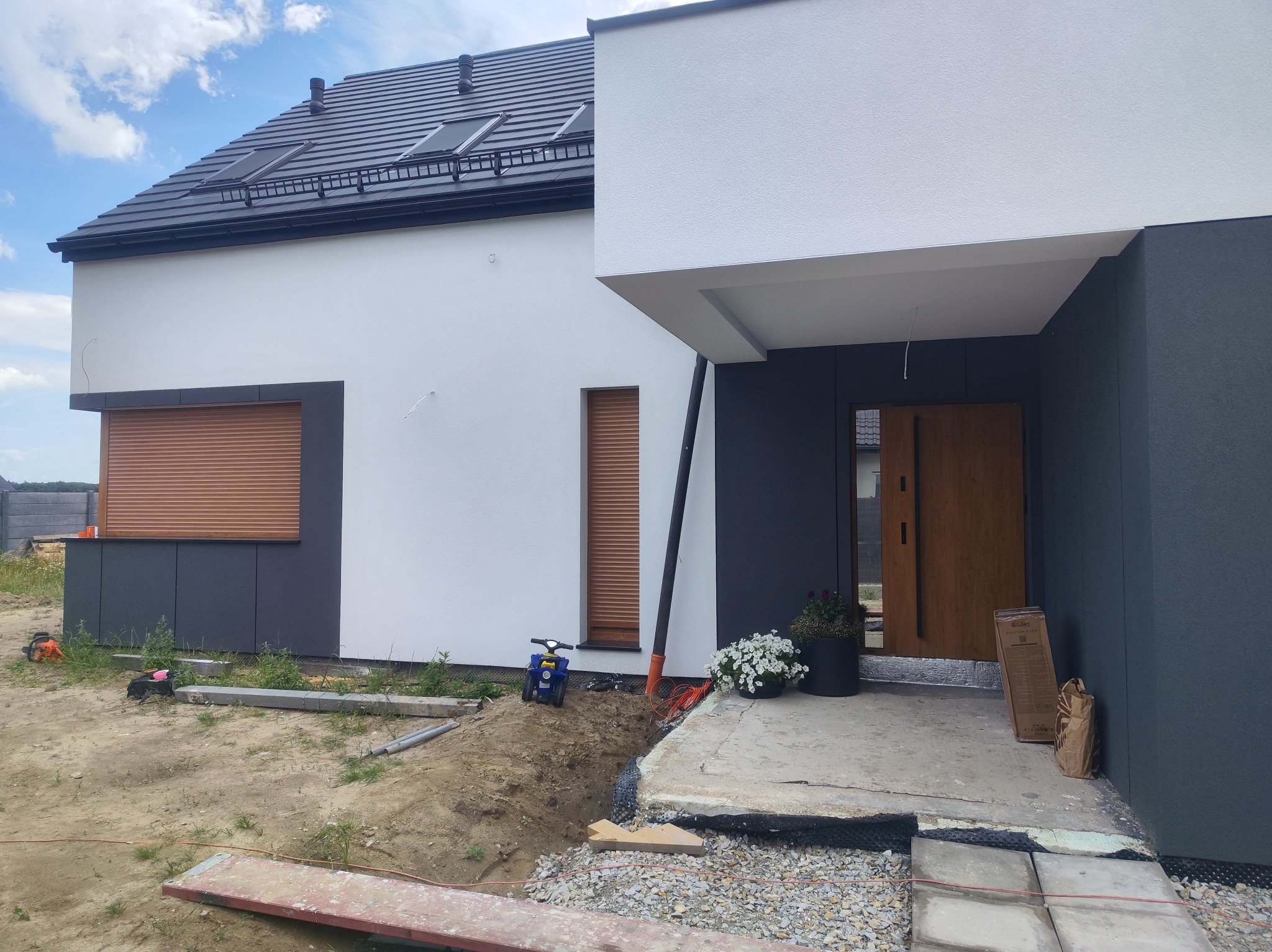 Nowoczesny dom jednorodzinny z białymi ścianami i grafitowymi elementami elewacji, drewnianymi roletami zewnętrznymi, czarnym dachem z oknami dachowymi, betonowym podjazdem w trakcie budowy...