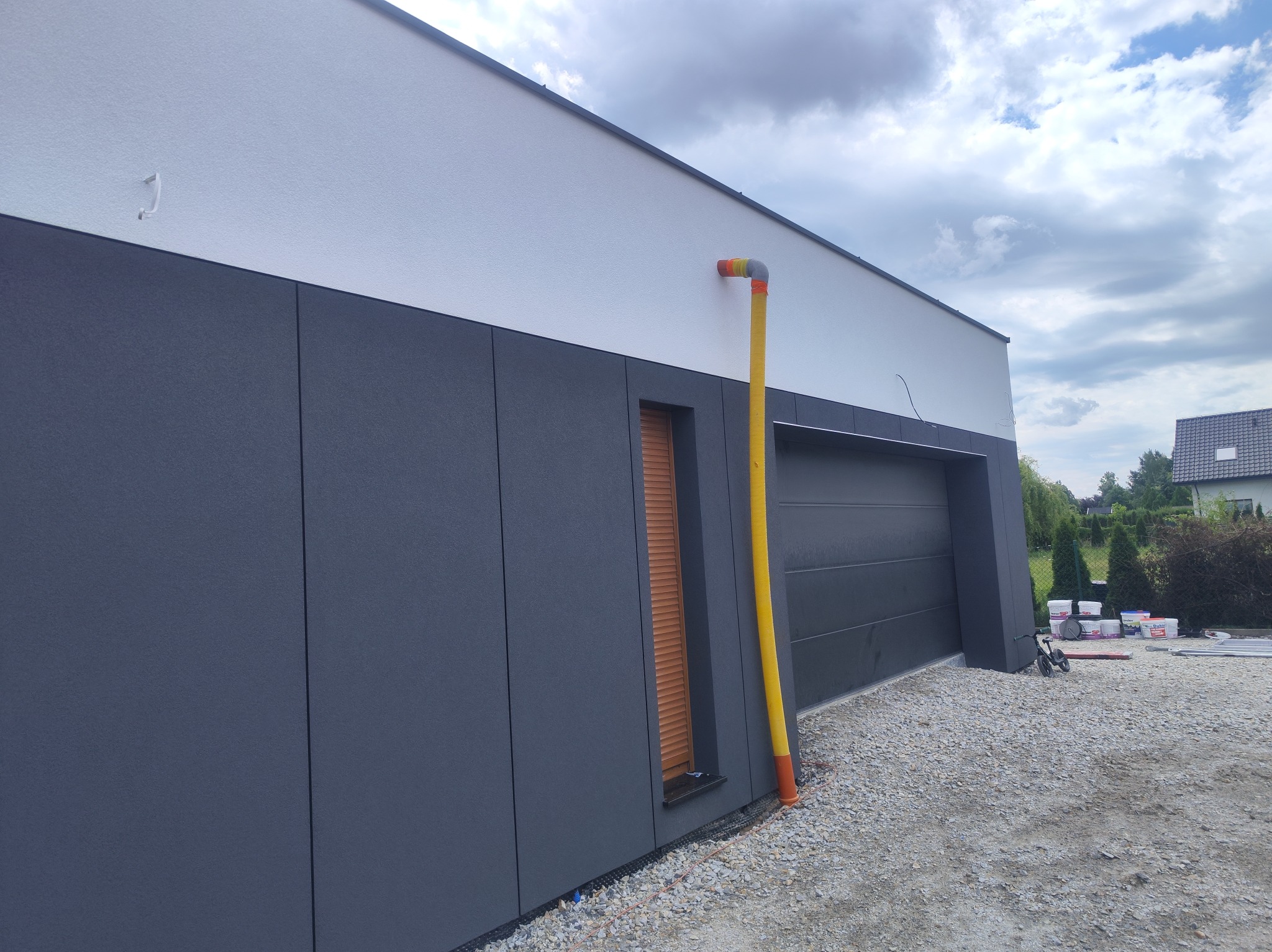 Dwukolorowa elewacja domu z szarymi panelami i białym tynkiem, widoczna żółta rura spustowa, okno z brązowymi żaluzjami i szara brama garażowa, teren wokół wysypany żwirem.