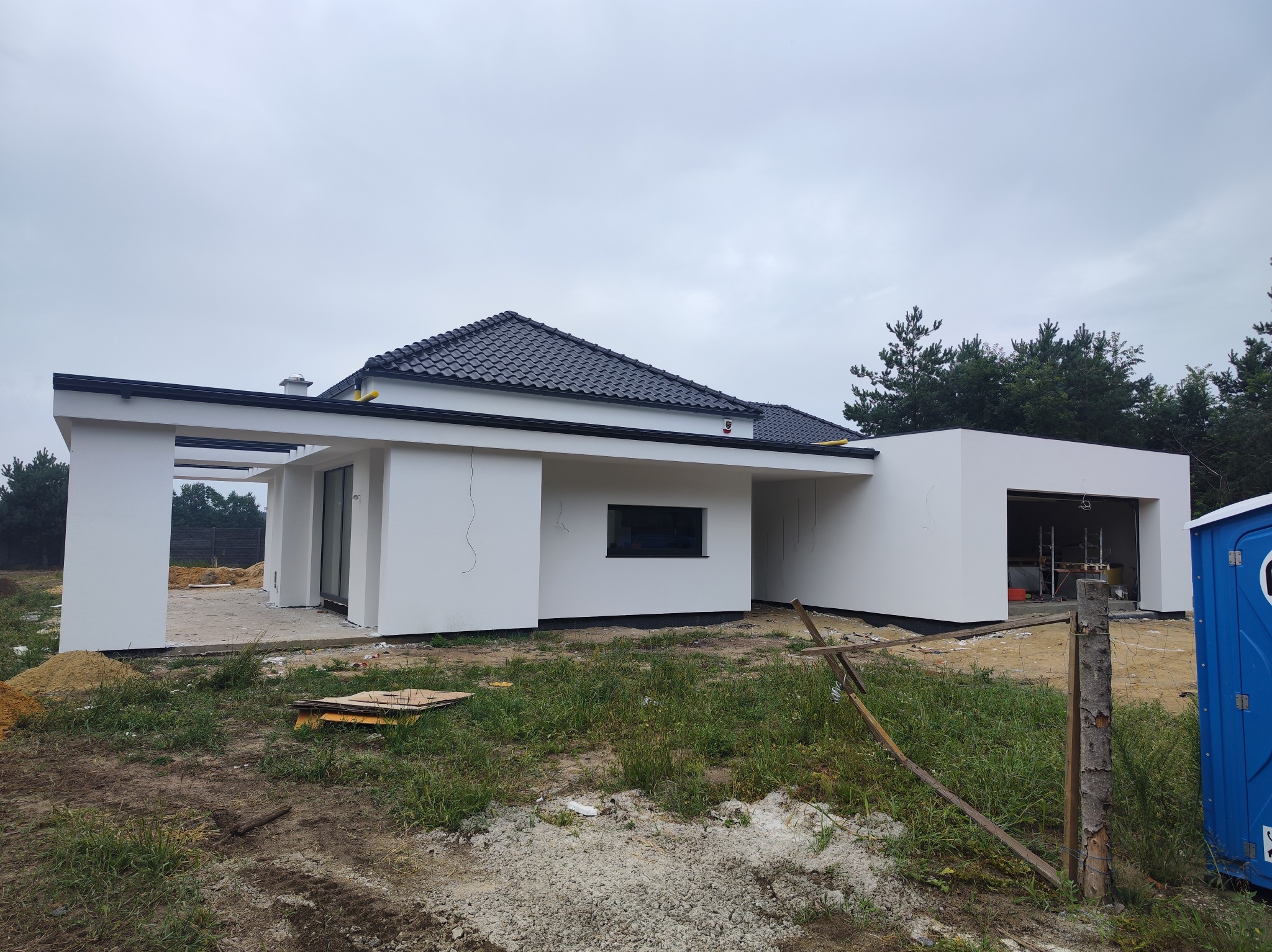 Nowoczesny dom jednorodzinny z białymi ścianami zewnętrznymi i ciemną dachówką, widoczny w trakcie prac wykończeniowych na zewnątrz, z fragmentami niezagospodarowanego terenu i przenośną toaletą.
