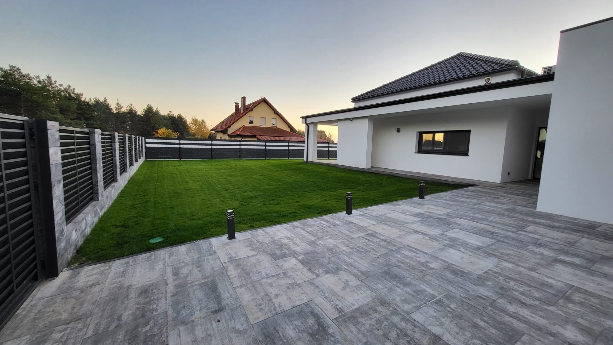 Nowoczesny dom jednorodzinny z zadbanym trawnikiem, szarą kostką brukową i ogrodzeniem panelowym w odcieniach szarości, widok na sąsiedni dom w tle.