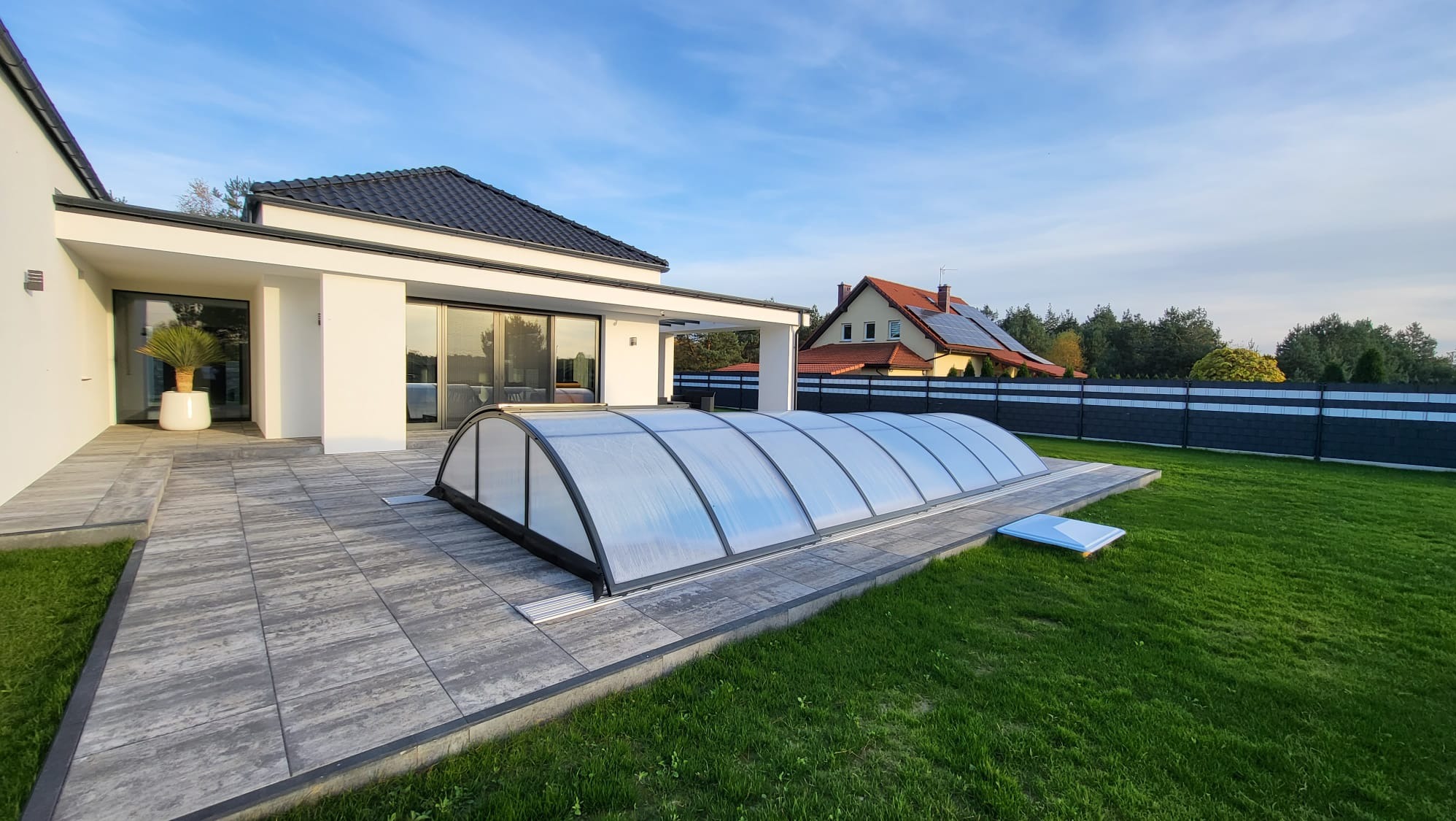 Zadaszenie basenowe z poliwęglanu na tarasie z szarych płyt betonowych przy nowoczesnym domu z białymi ścianami i czarnym dachem.