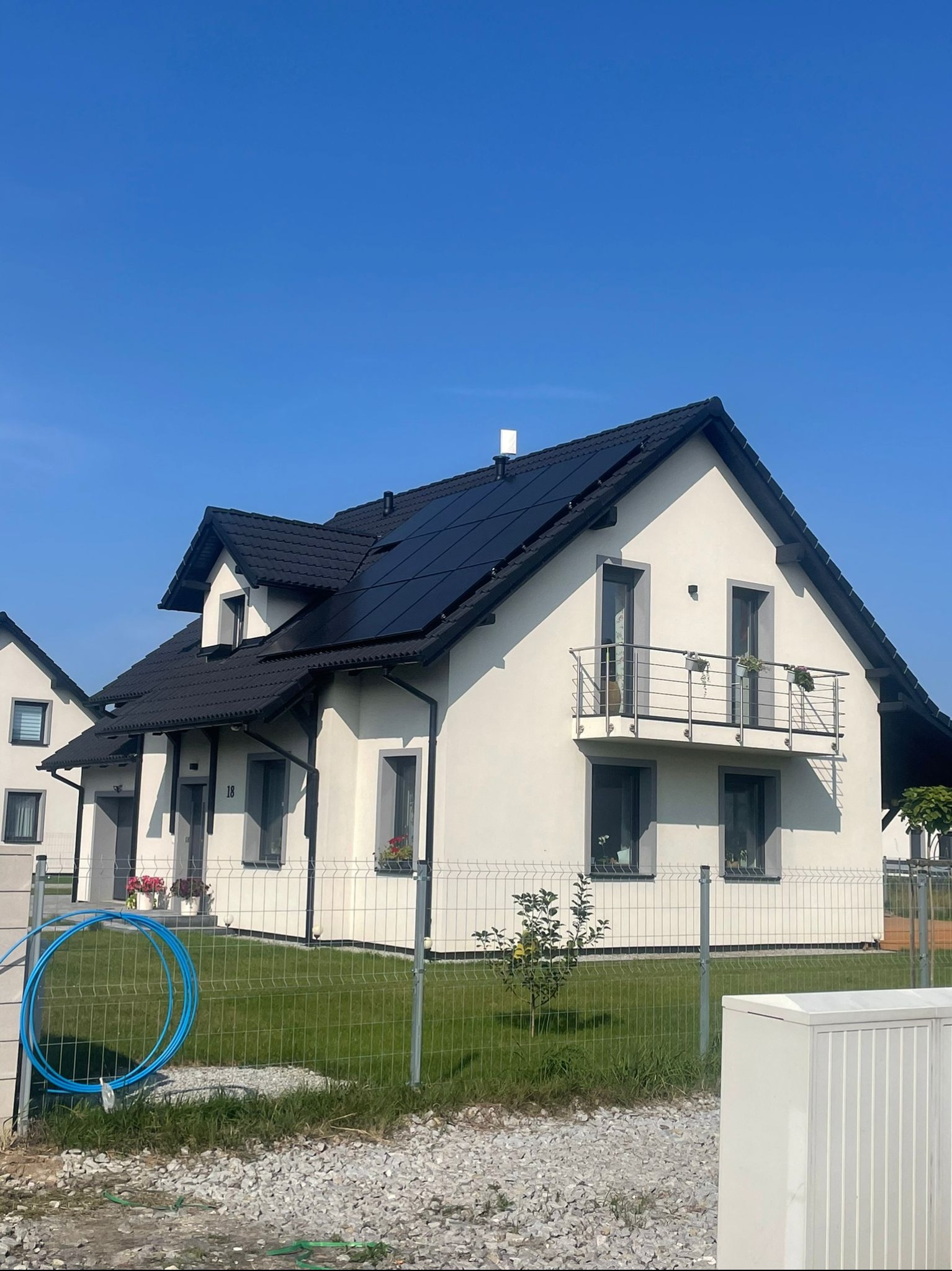 Nowoczesny dom jednorodzinny z balkonem i panelami słonecznymi na dachu, widoczny zza ogrodzenia panelowego, na tle błękitnego nieba.
