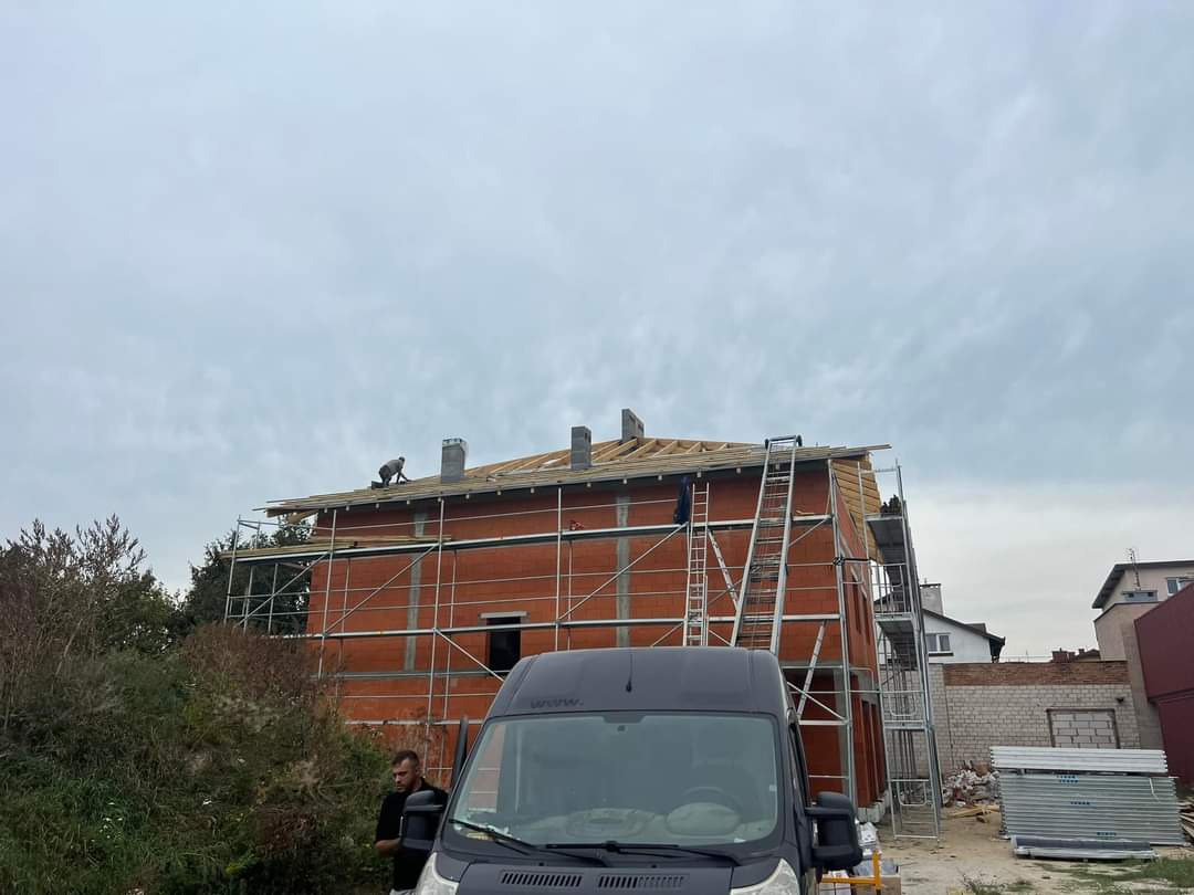 Budowa domu jednorodzinnego z widoczną konstrukcją dachu i rusztowaniem, dwóch pracowników na dachu oraz czarny samochód dostawczy na pierwszym planie.