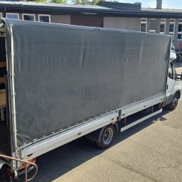 CAPY SPÓŁKA Z OGRANICZONĄ ODPOWIEDZIALNOŚCIĄ - Biały bus z otwartą plandeką, wewnątrz zapakowany stół. Widoczny fragment budynku w tle. Transport mebli.