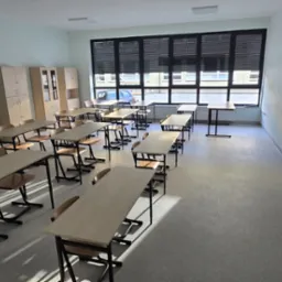 Jasna klasa z rzędami ławek i krzeseł, szafy w rogu i duże okno z żaluzjami. Świeżo pomalowane ściany i czysta podłoga.