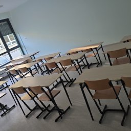 CAPY SPÓŁKA Z OGRANICZONĄ ODPOWIEDZIALNOŚCIĄ - Ustawione rzędy jasnych biurek i krzeseł z czarnymi nogami w pustej klasie, widok z boku, okno z żaluzjami w tle.