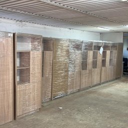 CAPY SPÓŁKA Z OGRANICZONĄ ODPOWIEDZIALNOŚCIĄ - Szafy w folii stretch, gotowe do transportu, ustawione w rzędzie w magazynie. Widoczna opona samochodowa i biała tablica z notatkami.