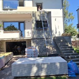 CAPY SPÓŁKA Z OGRANICZONĄ ODPOWIEDZIALNOŚCIĄ - Dwupiętrowy dom w trakcie remontu elewacji. Na pierwszym piętrze widoczne okna, na drugim pracownik na drabinie. Na dole składowane płyty gipsowe i worki z zaprawą.