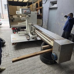 CAPY SPÓŁKA Z OGRANICZONĄ ODPOWIEDZIALNOŚCIĄ - Załadunek maszyny do transportu busem. Sprzęt zabezpieczony na oponach i drewnianych belkach. Osoba w oddali. Tył samochodu dostawczego z otwartą burtą.