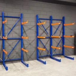 Dwa niebieskie regały magazynowe z pomarańczowymi wspornikami na szarej ścianie z cegieł. Obok sprężarka powietrza. Na podłodze poziomica.