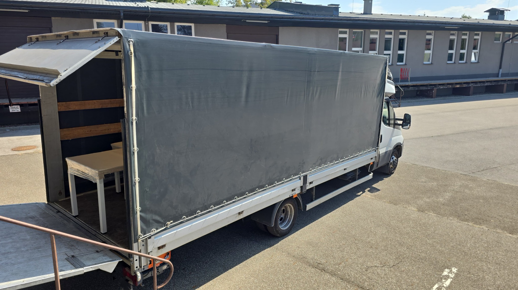 Biały bus z otwartą plandeką, wewnątrz zapakowany stół. Widoczny fragment budynku w tle. Transport mebli.