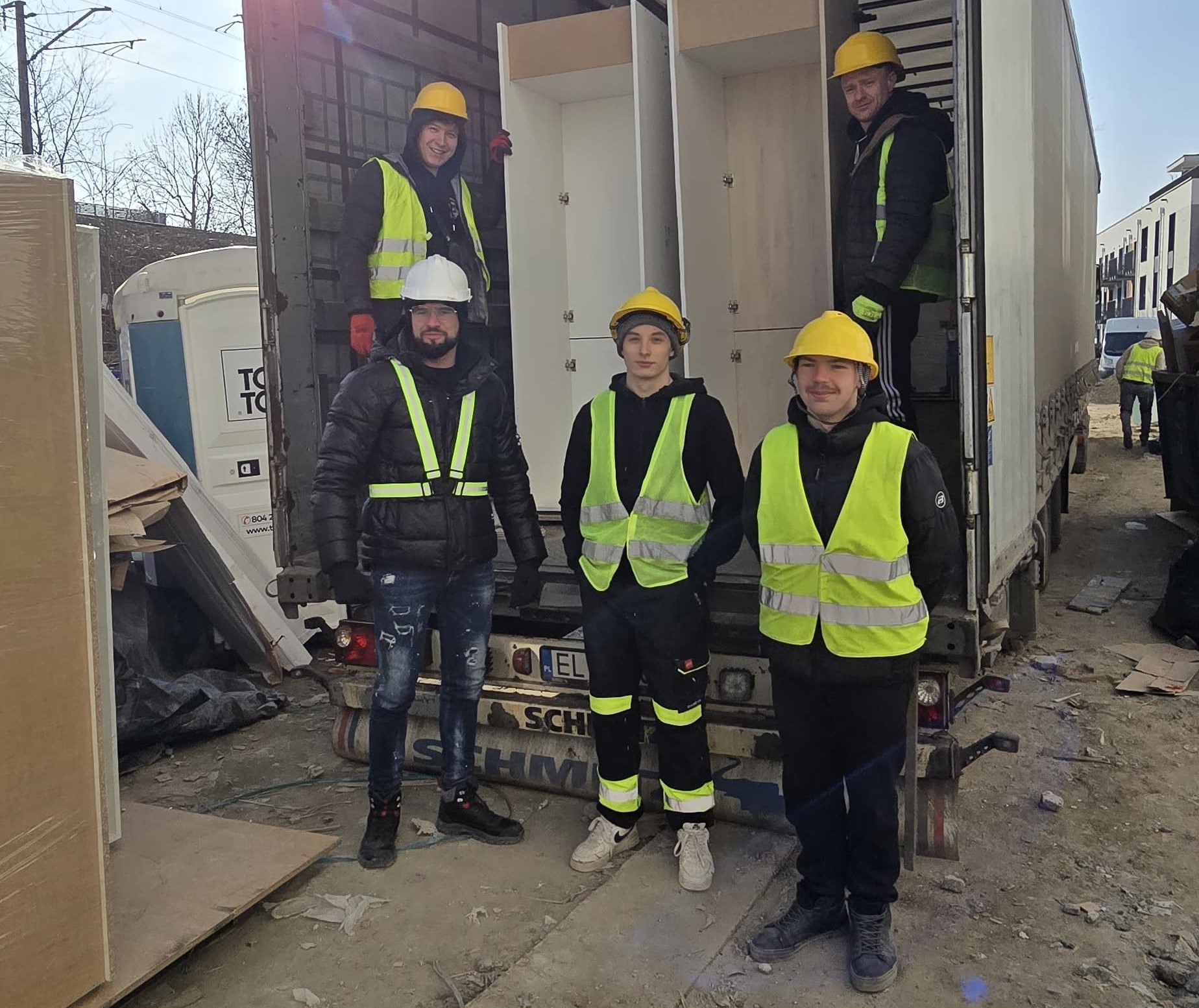 Ekipa w żółtych kamizelkach i kaskach rozładowuje białe szafy z ciężarówki na placu budowy. Widoczna przenośna toaleta i budynki w tle.