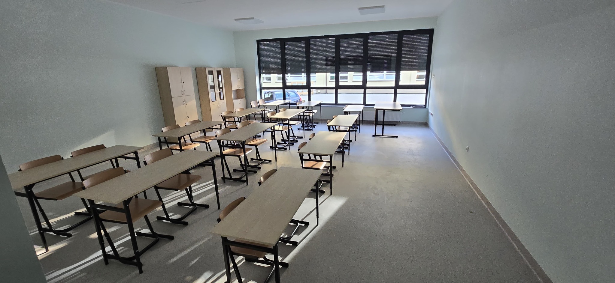 Jasna klasa z rzędami ławek i krzeseł, szafy w rogu i duże okno z żaluzjami. Świeżo pomalowane ściany i czysta podłoga.