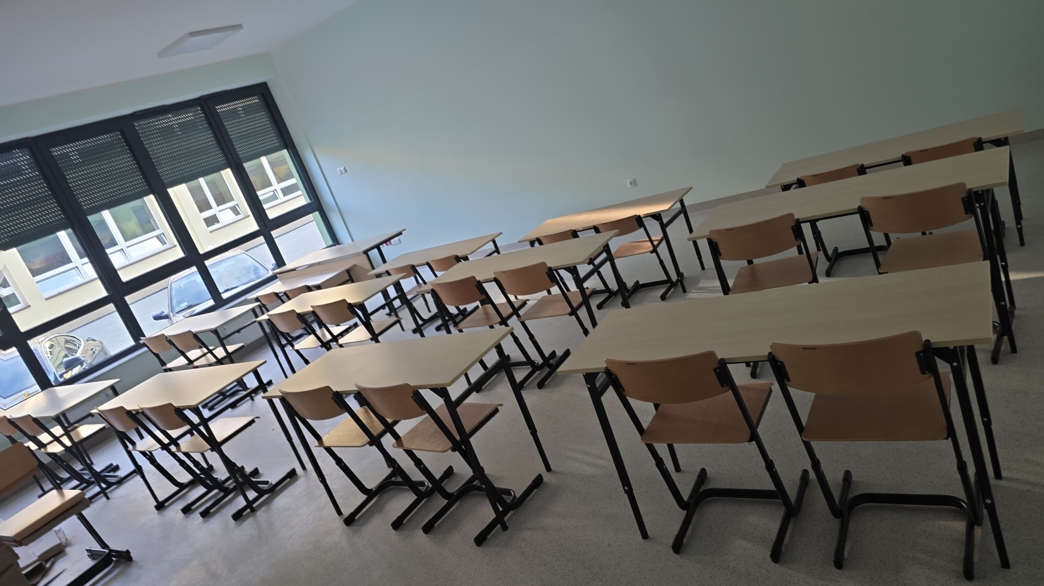 Ustawione rzędy jasnych biurek i krzeseł z czarnymi nogami w pustej klasie, widok z boku, okno z żaluzjami w tle.