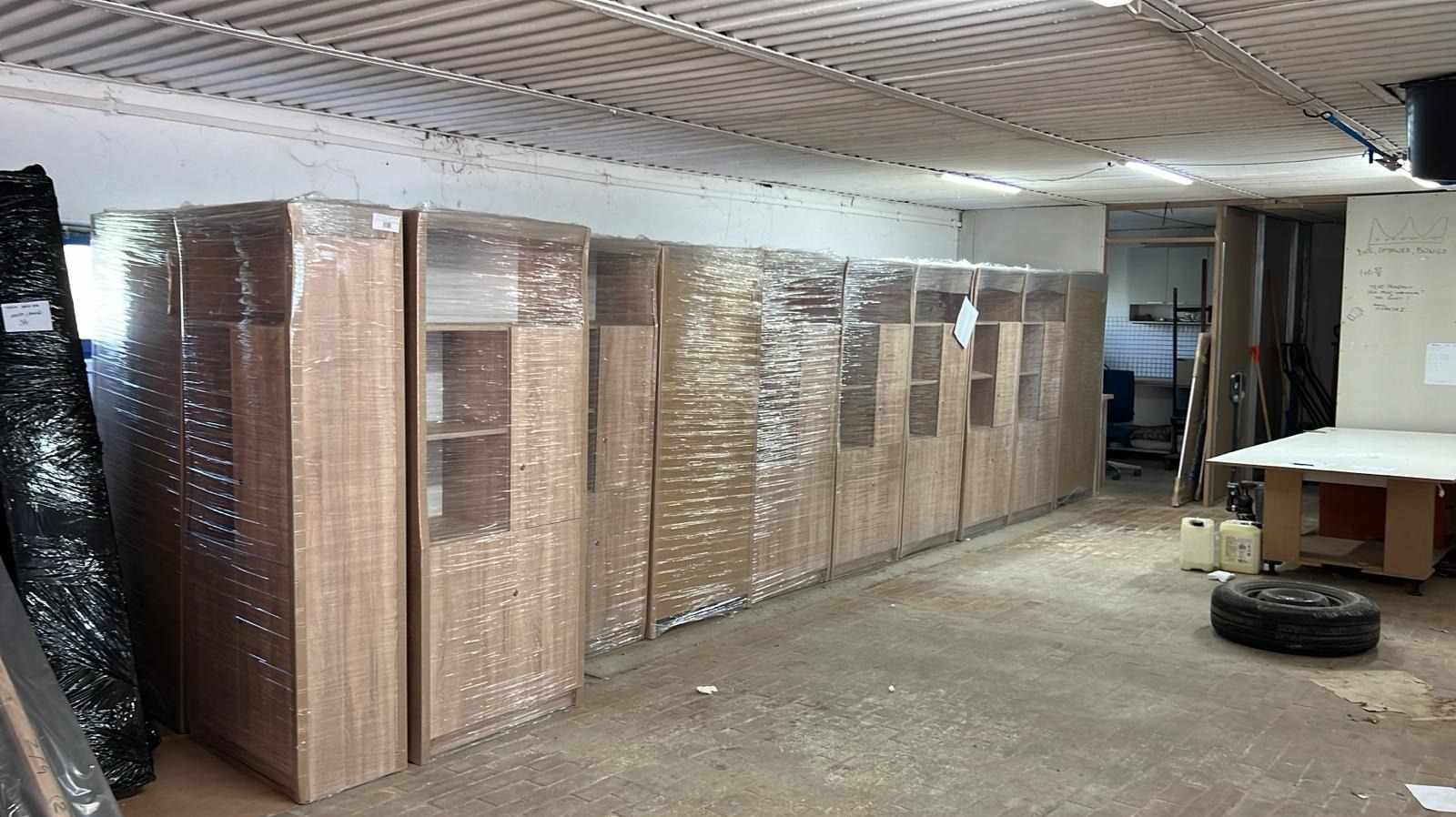 Szafy w folii stretch, gotowe do transportu, ustawione w rzędzie w magazynie. Widoczna opona samochodowa i biała tablica z notatkami.