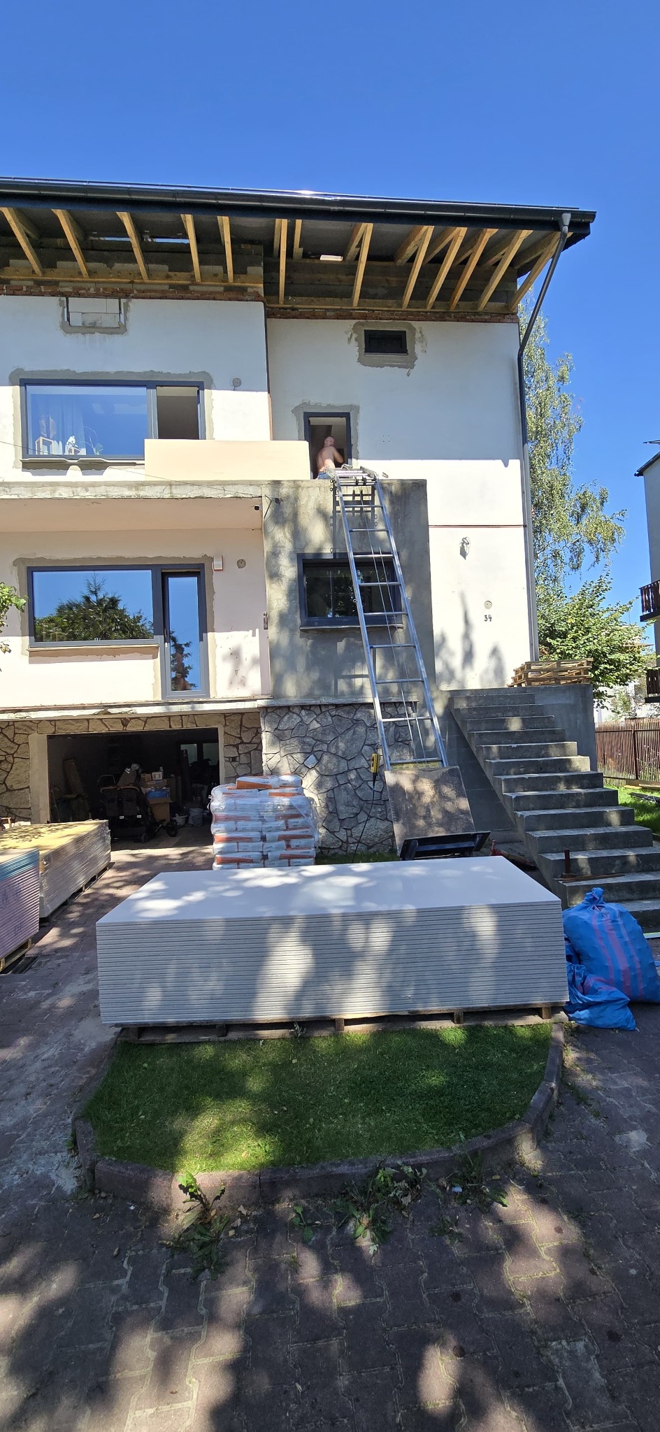 Dwupiętrowy dom w trakcie remontu elewacji. Na pierwszym piętrze widoczne okna, na drugim pracownik na drabinie. Na dole składowane płyty gipsowe i worki z zaprawą.