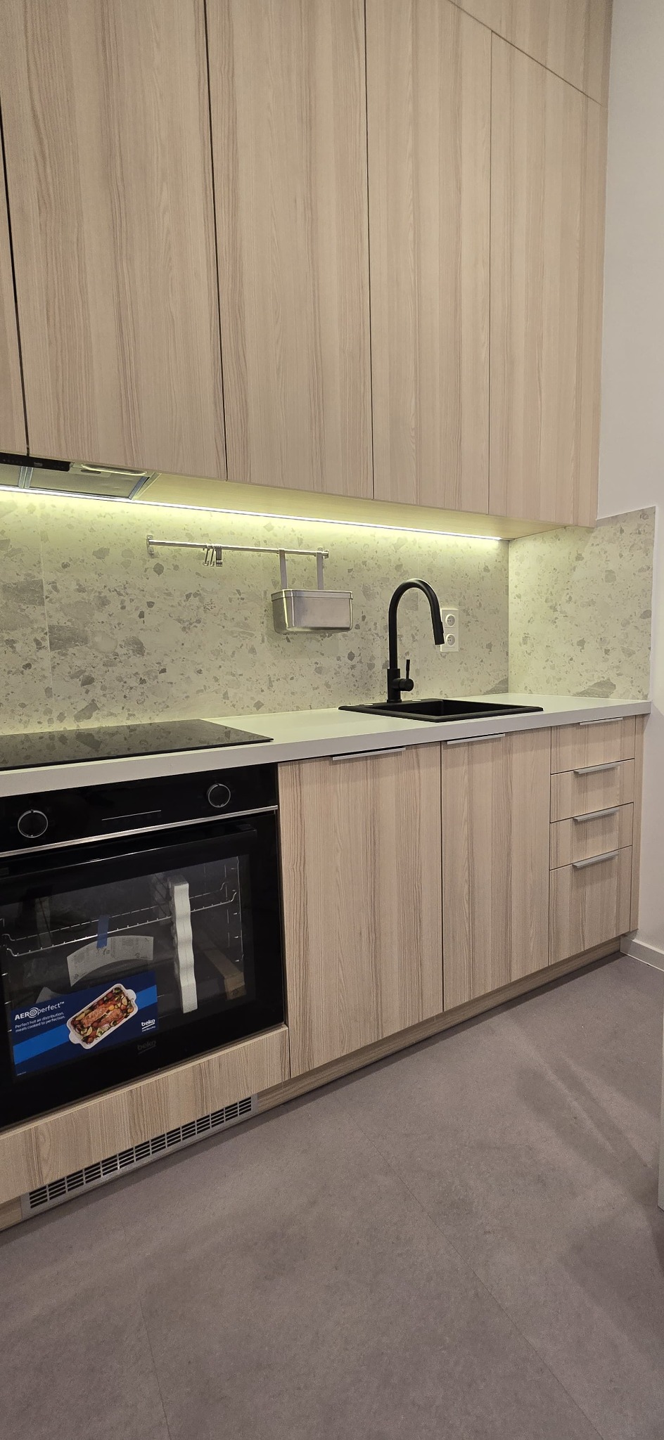 Nowoczesna kuchnia z drewnopodobnymi frontami, czarną armaturą i kamiennym blatem. Oświetlenie LED podkreśla minimalistyczny design. Widoczny piekarnik Beko.