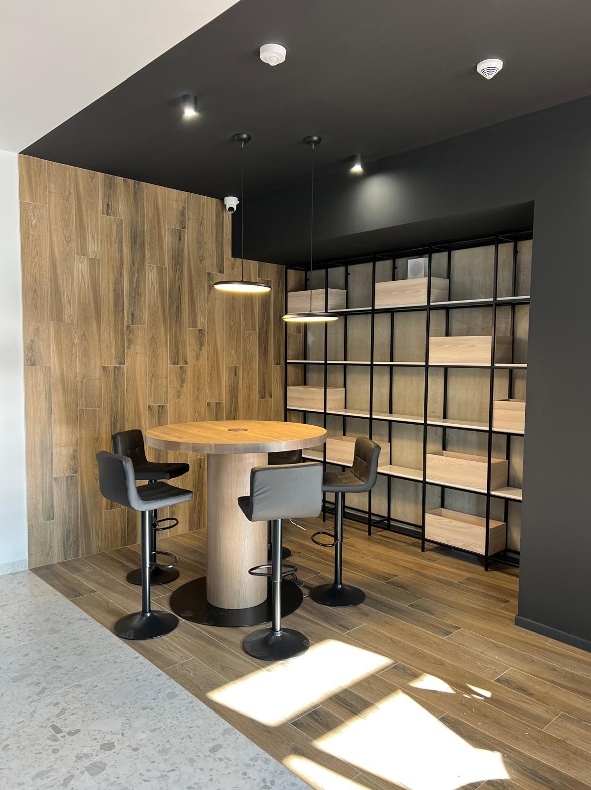 Stylowe biuro z okrągłym stołem barowym i czterema hokerami, drewnianą ścianą oraz regałem z półkami. Oświetlenie punktowe i wiszące lampy dodają nowoczesnego charakteru.