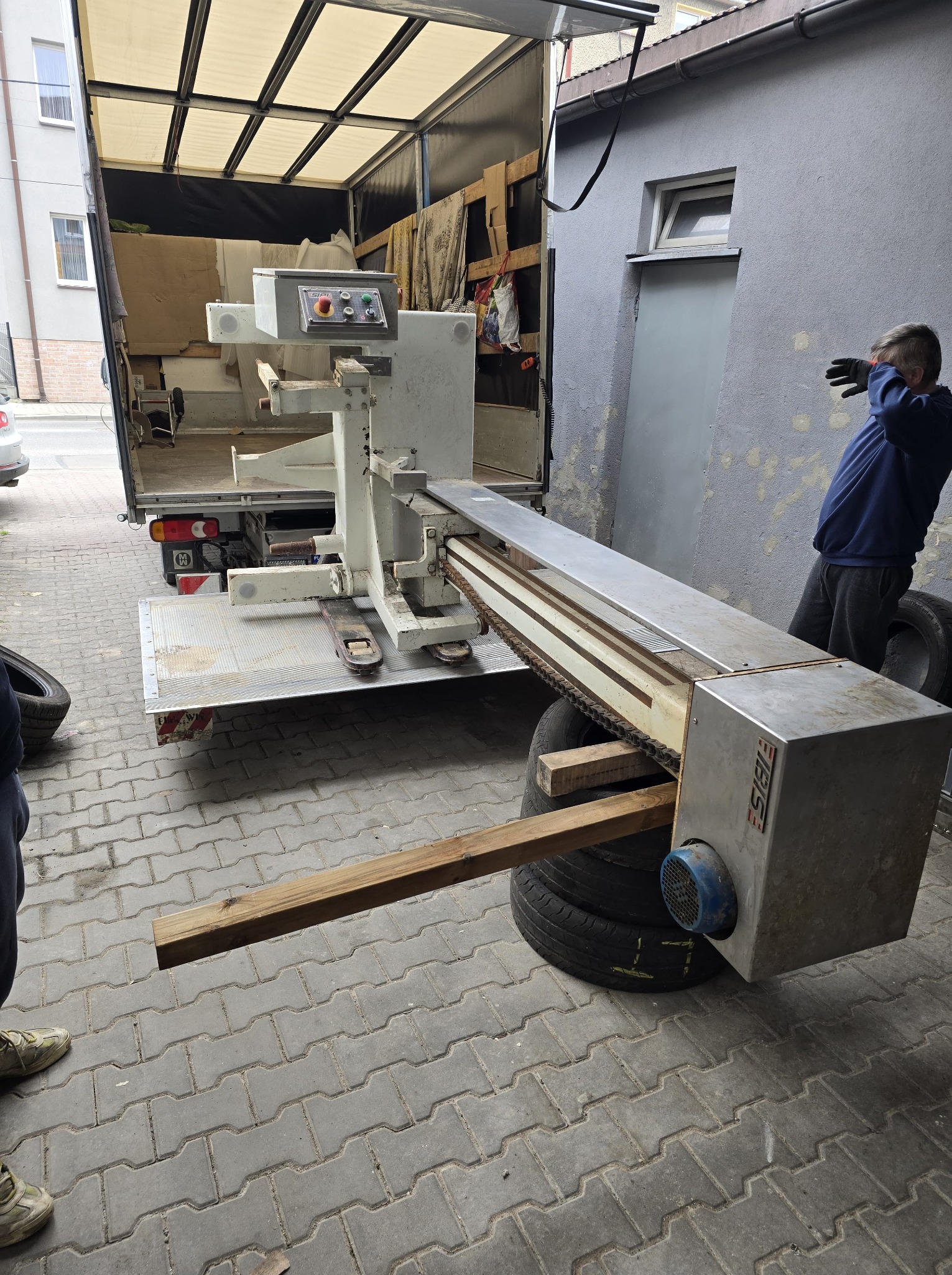 Załadunek maszyny do transportu busem. Sprzęt zabezpieczony na oponach i drewnianych belkach. Osoba w oddali. Tył samochodu dostawczego z otwartą burtą.