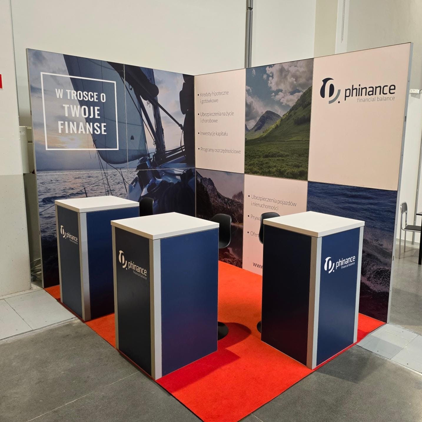 Stoisko targowe Phinance z logo, grafiką żaglówki i gór. Trzy biało-niebieskie lady na czerwonej wykładzinie. Prezentacja oferty finansowej.