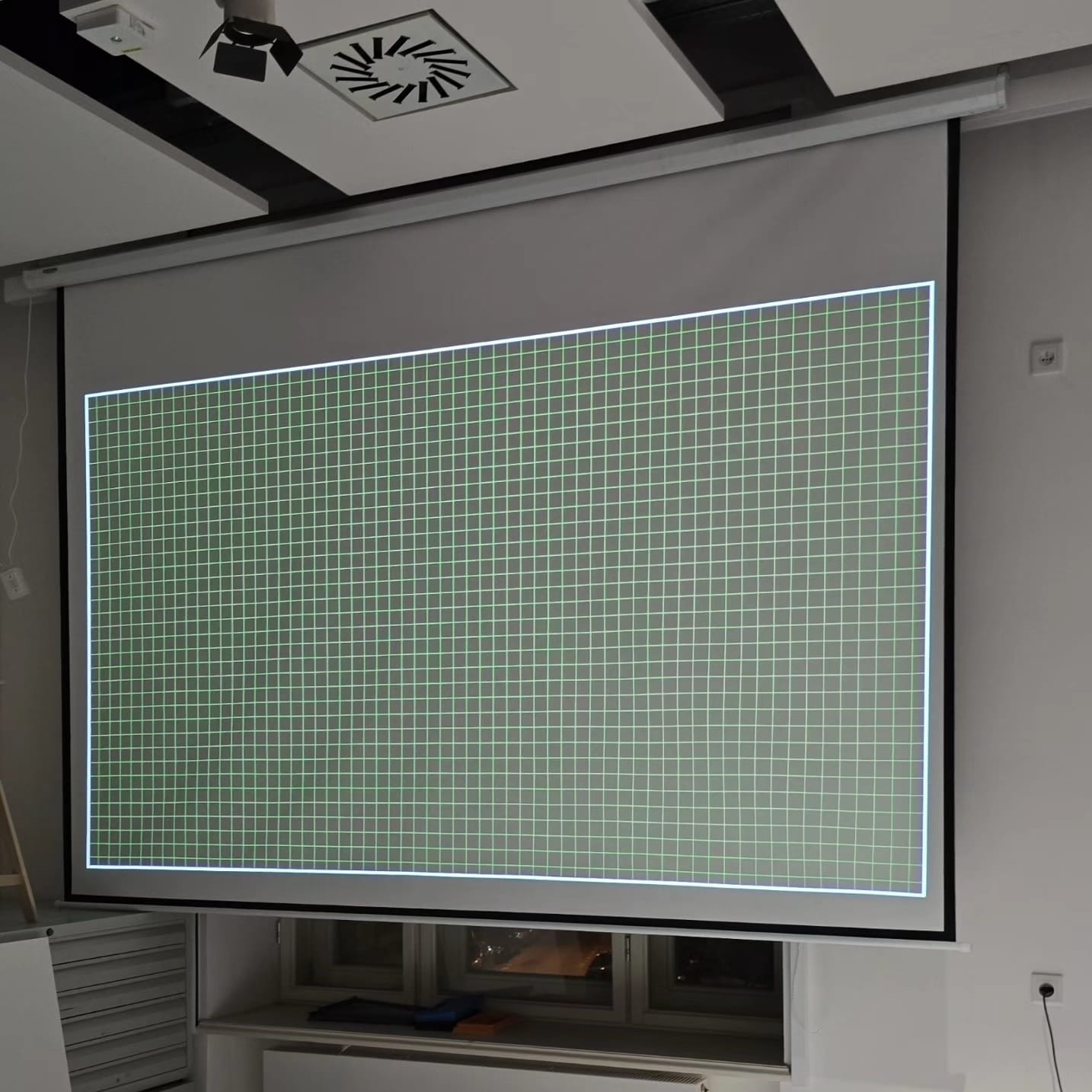 Zamontowany ekran projekcyjny z wyświetlaną siatką kalibracyjną w jasnym biurze. Widoczne okno pod ekranem i oświetlenie sufitowe.