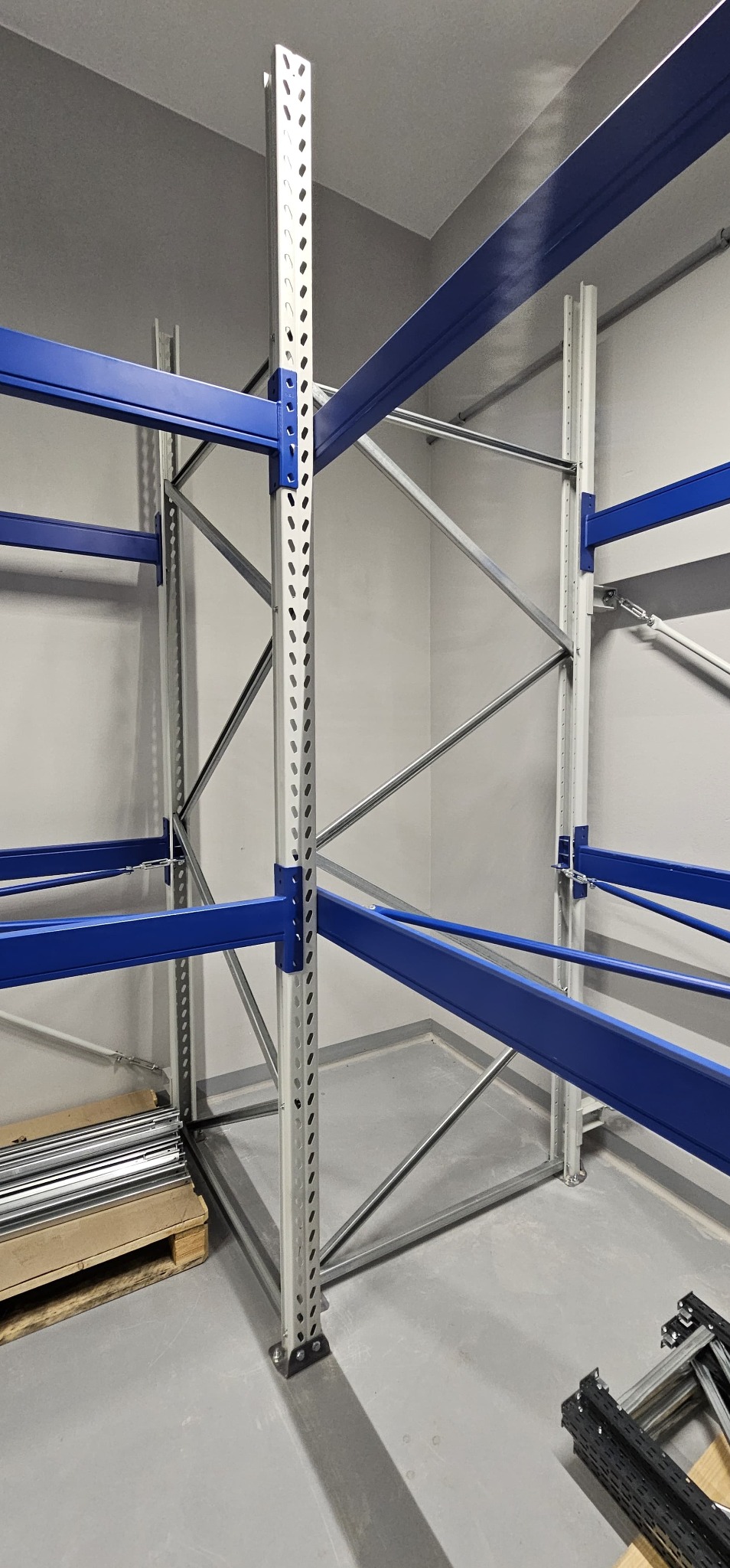 Montaż regału magazynowego: stalowe słupy z perforacją, niebieskie belki poprzeczne, szare ściany i podłoga, elementy do montażu na palecie, surowy minimalizm.
