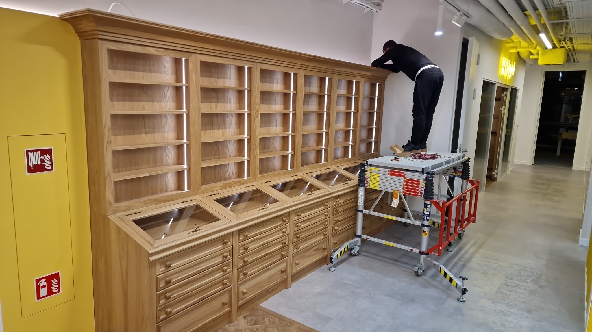Stolarz na rusztowaniu montuje regał z szufladami i przeszklonymi gablotami w biurze. Żółta ściana z hydrantem po lewej, korytarz z neonem w tle.