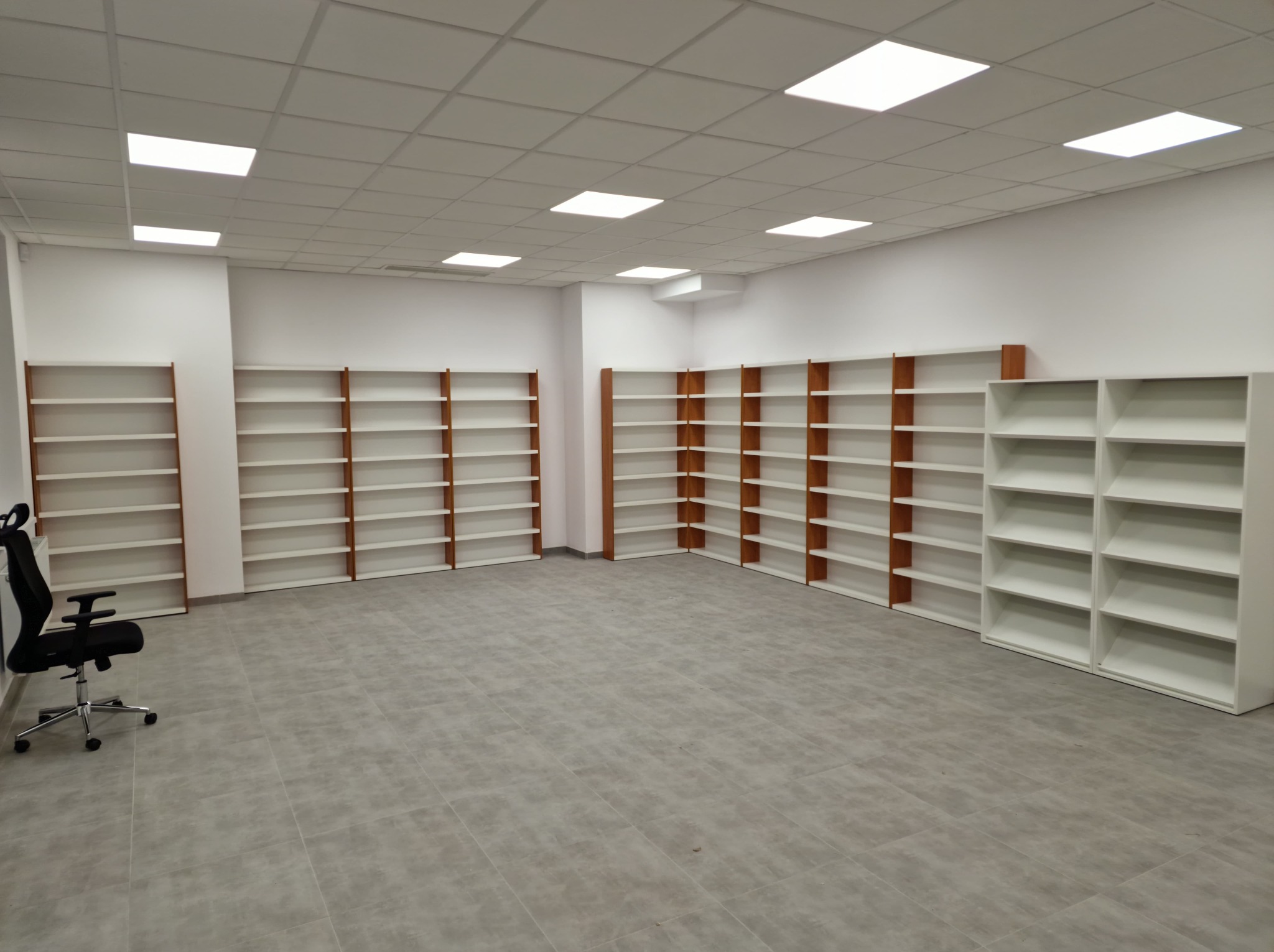 Jasne biuro z nowymi, białymi regałami z brązowymi akcentami. Krzesło biurowe na kółkach stoi z boku, na szarej podłodze z płytek. Oświetlenie sufitowe.