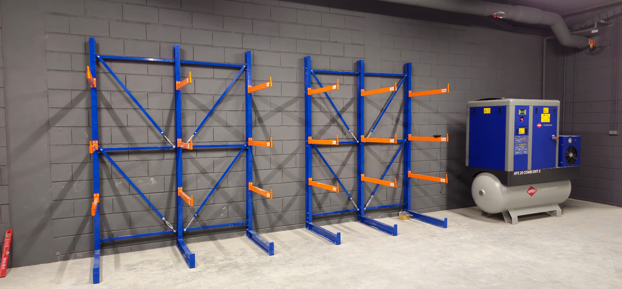 Dwa niebieskie regały magazynowe z pomarańczowymi wspornikami na szarej ścianie z cegieł. Obok sprężarka powietrza. Na podłodze poziomica.