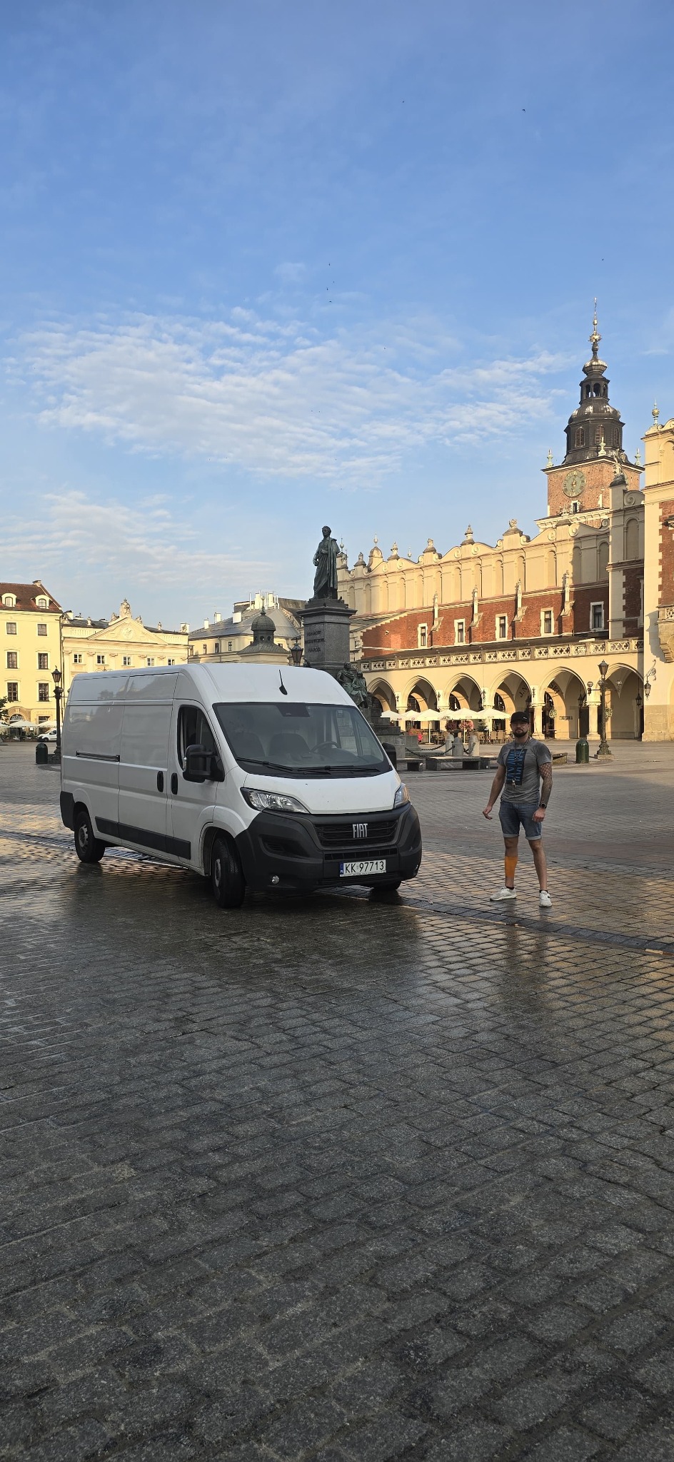 Biały furgon Fiat z numerem rejestracyjnym KK 97713 na tle Sukiennic w Krakowie, obok stoi mężczyzna. Usługi transportowe w zabytkowej scenerii.