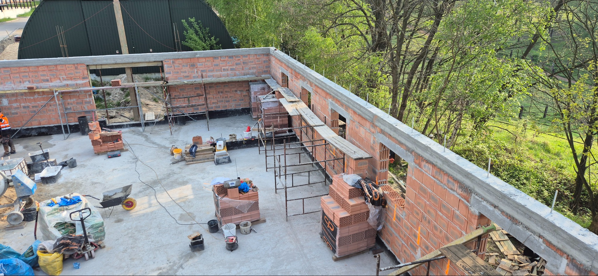 Wysoki kąt: budowa domu z czerwonej cegły, widoczne rusztowania, materiały budowlane i pracownik w kamizelce odblaskowej. Tło stanowią drzewa.