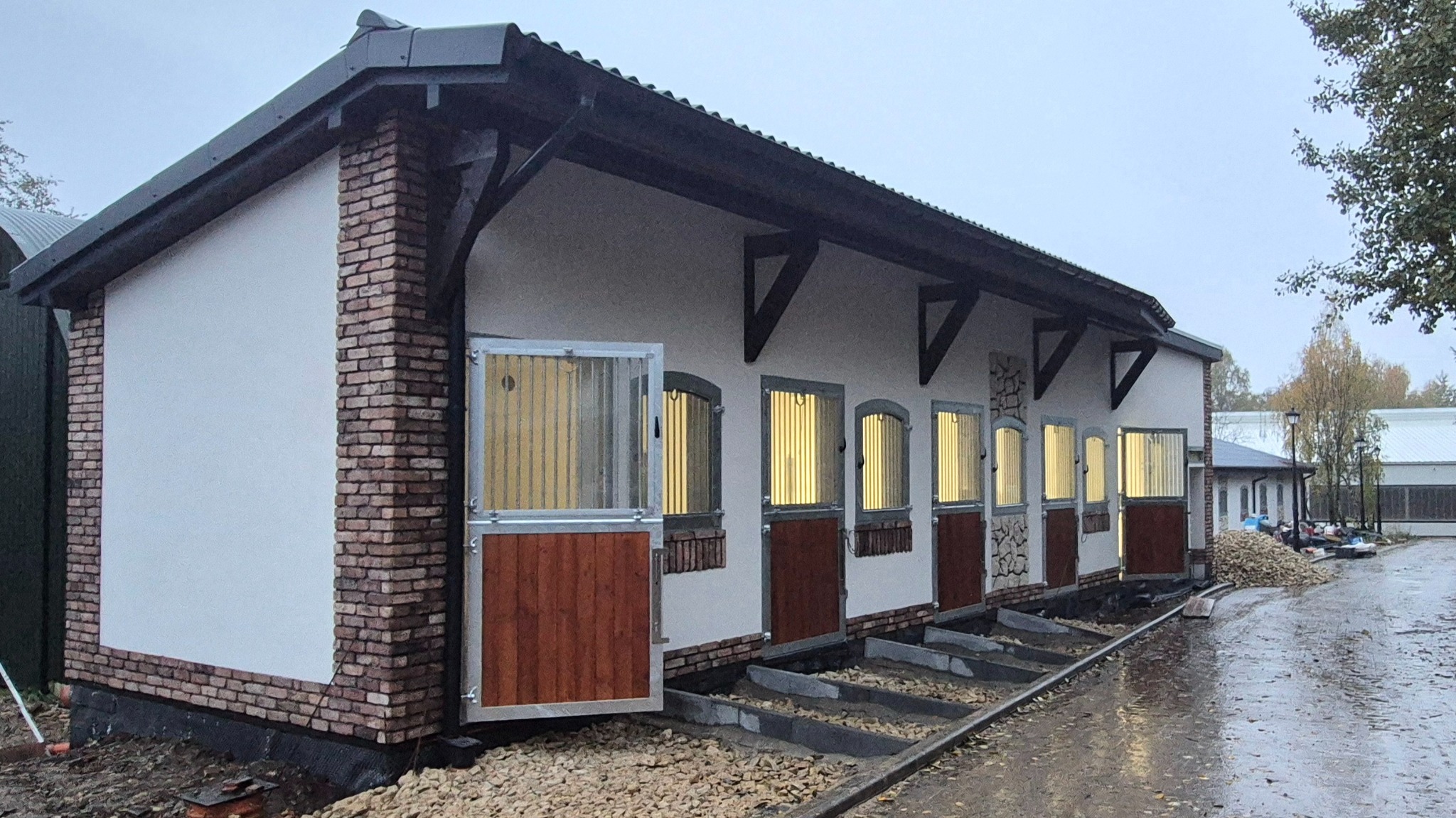 Nowo wybudowany budynek stajni z ceglanymi detalami, otwartymi drzwiami i oknami z kratami, oświetlony od wewnątrz, na zewnątrz widoczne kamienie i mokra nawierzchnia.