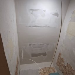 Usługi remontowo-budowlane Paweł Biel - Fragment wnętrza w trakcie remontu: zagruntowany sufit z widocznymi śladami napraw, fragmenty starej tapety na ścianach, przewody elektryczne i lampa oświetlająca przestrzeń.