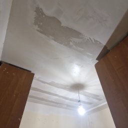Usługi remontowo-budowlane Paweł Biel - Niedokończona gładź na suficie w pokoju, widoczne fragmenty surowego tynku i fragment szafki. Żarówka w tle. Prace remontowe w trakcie.