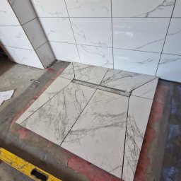 Usługi remontowo-budowlane Paweł Biel - Układanie marmurowych płytek w łazience, widoczny odpływ liniowy. Poziomica leży na podłodze. Płytki na ścianie w trakcie montażu, szare fugi.