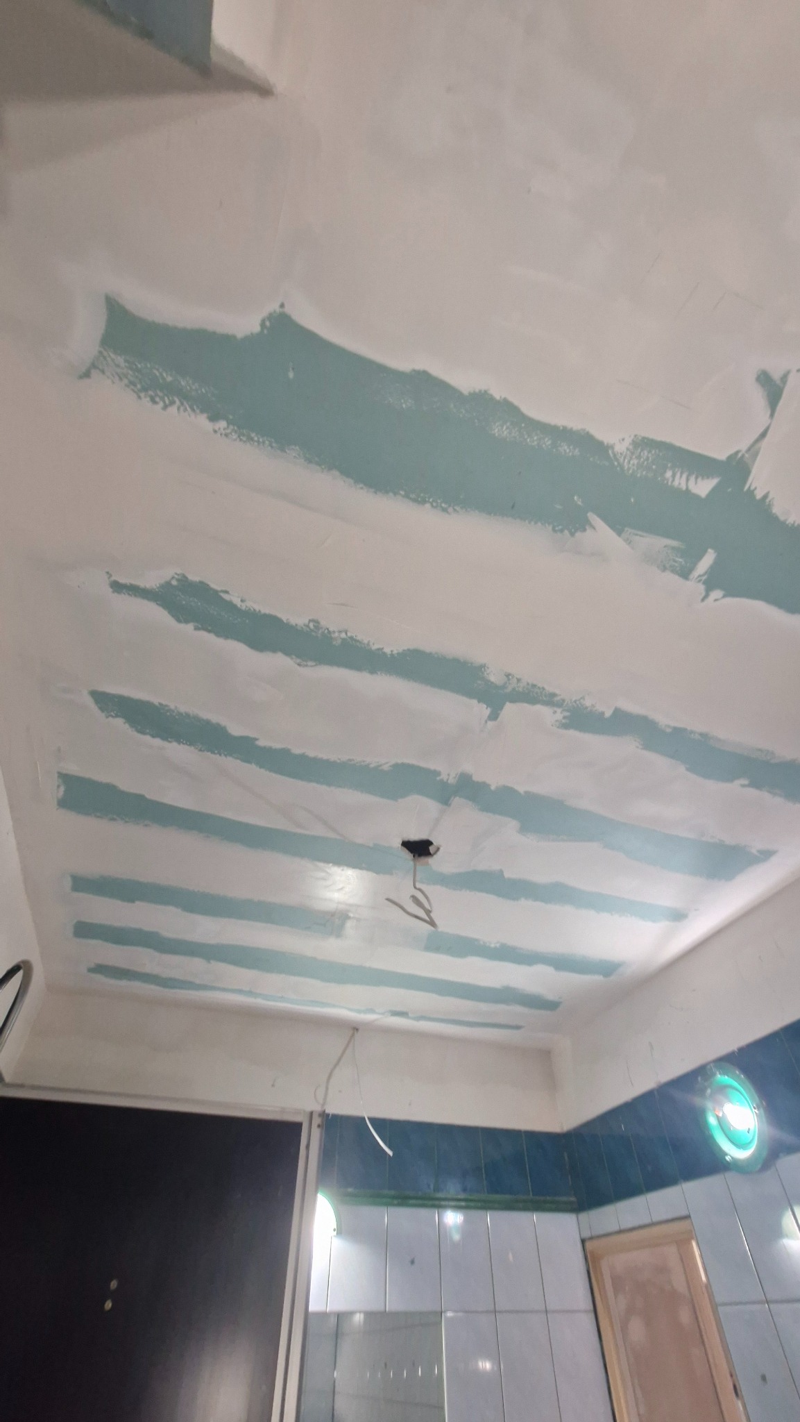 Niedokończone gładzie na suficie w łazience, widoczne pasy szpachli w kolorze niebieskim, przygotowanie do malowania. W tle glazura i fragment drzwi.