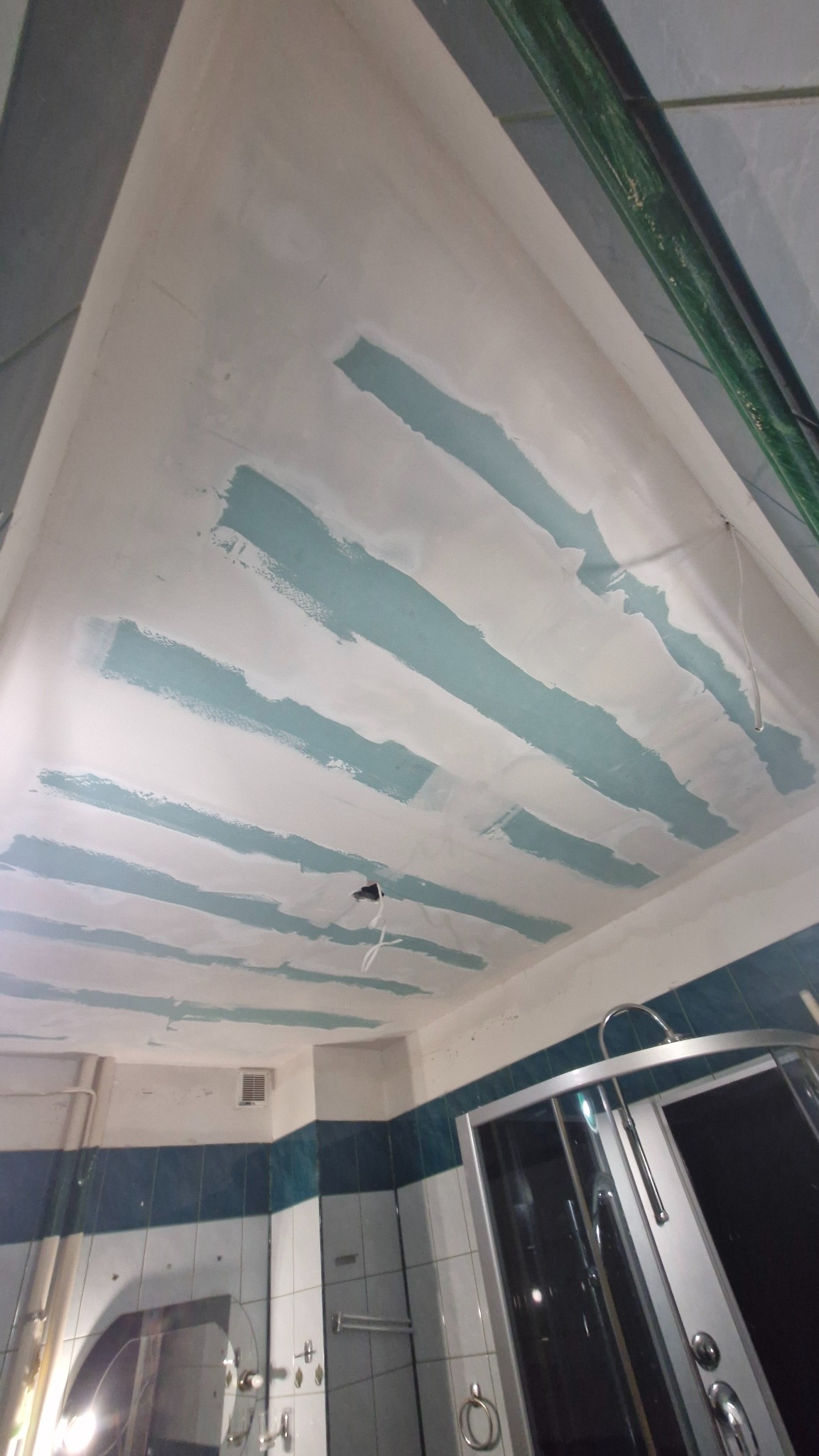 Nisko kątowe ujęcie sufitu łazienki w trakcie gładzenia, z widocznymi pasami nałożonej gładzi i fragmentem kabiny prysznicowej w tle.