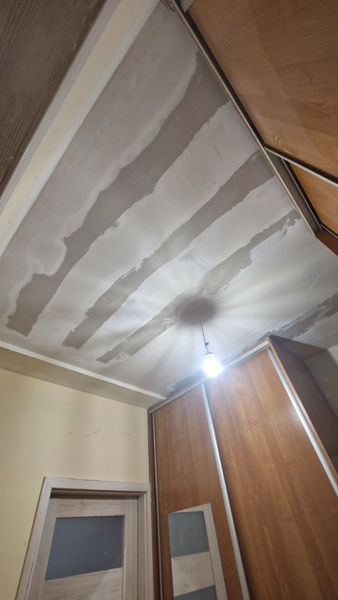 Nisko kątowe ujęcie sufitu w trakcie prac wykończeniowych, widoczne pasy świeżo nałożonej gładzi, oświetlone żarówką. W tle fragment szafy i drzwi.