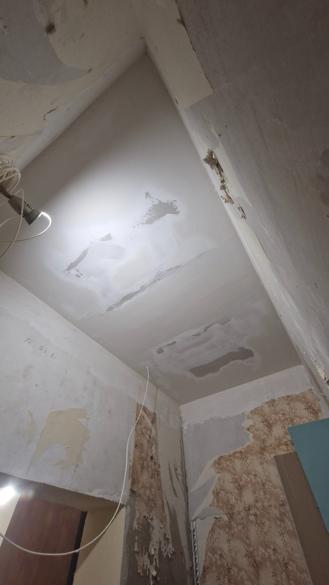 Pokój w trakcie remontu: ściany z resztkami tapet, sufit z widocznymi plamami i miejscami po naprawach, oświetlenie robocze, przygotowanie do malowania.