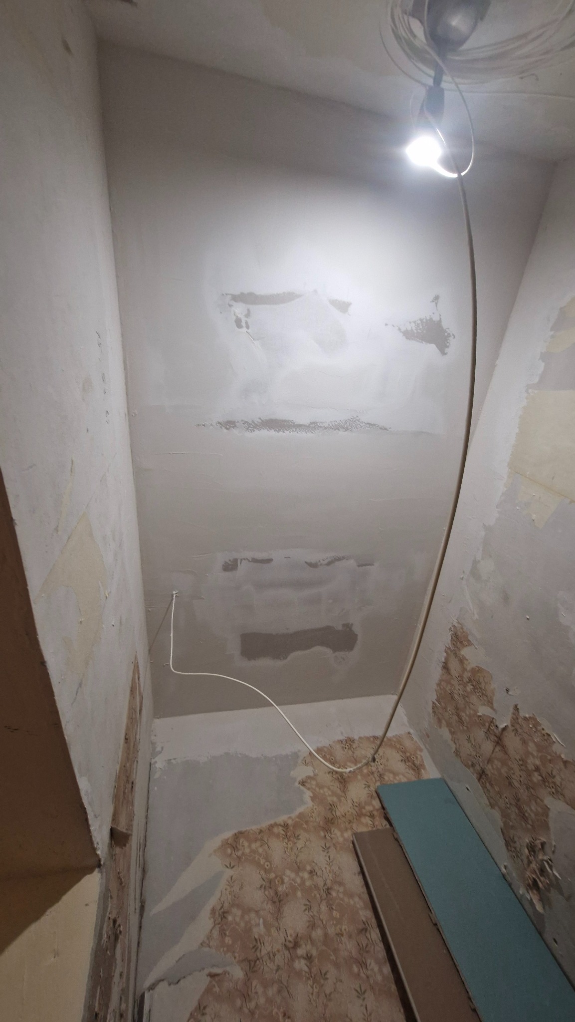Fragment wnętrza w trakcie remontu: zagruntowany sufit z widocznymi śladami napraw, fragmenty starej tapety na ścianach, przewody elektryczne i lampa oświetlająca przestrzeń.