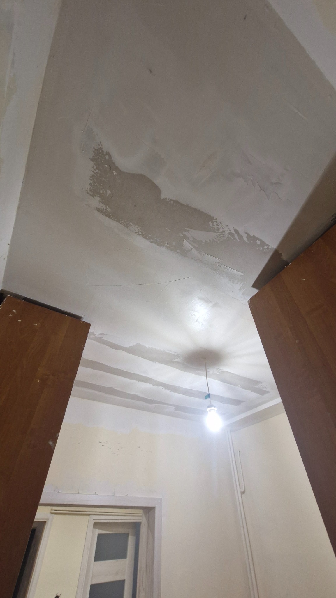 Niedokończona gładź na suficie w pokoju, widoczne fragmenty surowego tynku i fragment szafki. Żarówka w tle. Prace remontowe w trakcie.