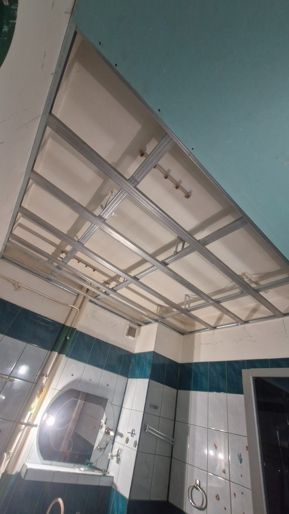 Stelaż sufitu podwieszanego z płyt karton-gips w łazience w trakcie montażu. Widoczne profile metalowe, częściowo zamontowane płyty i instalacje.