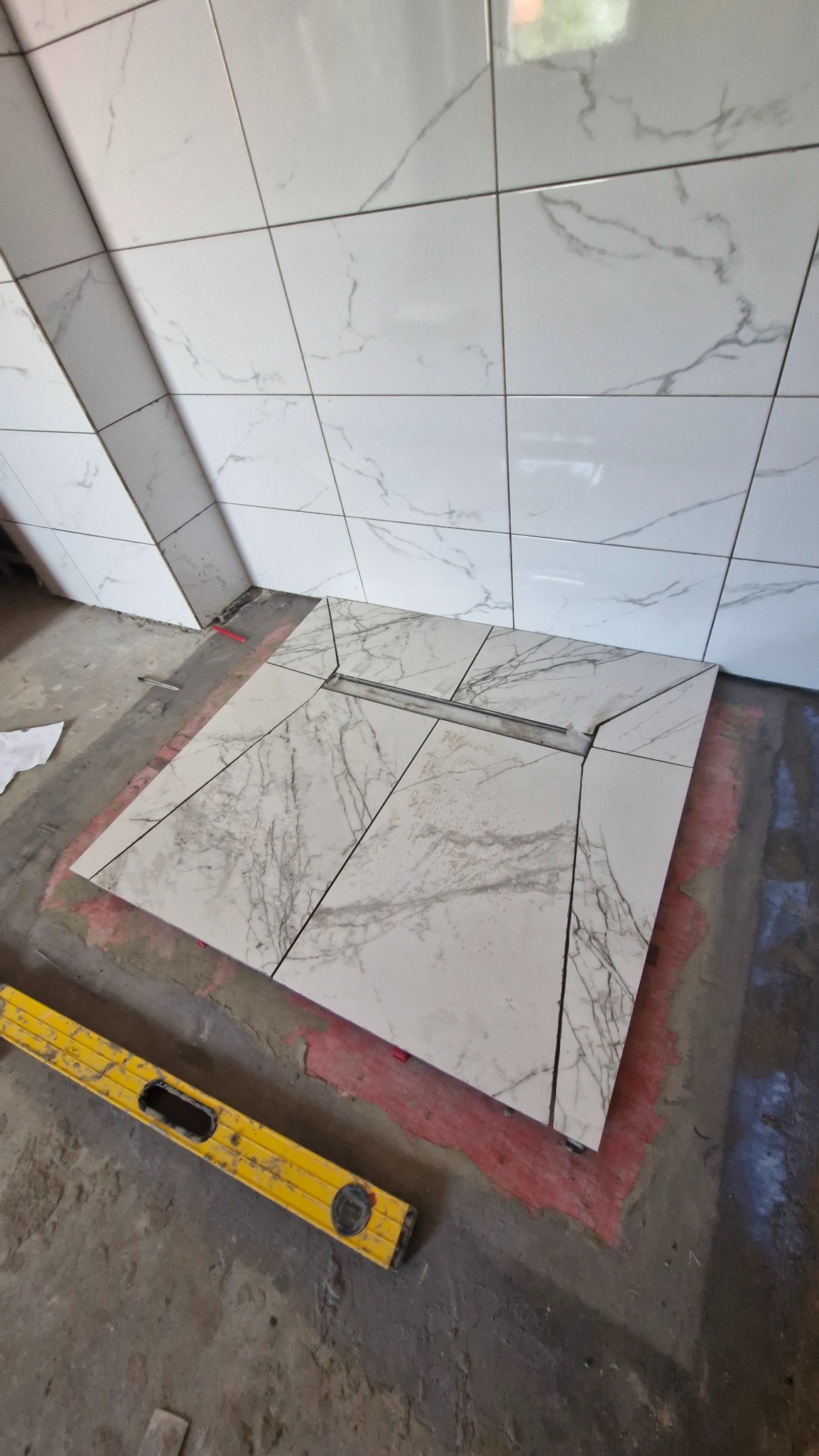 Układanie marmurowych płytek w łazience, widoczny odpływ liniowy. Poziomica leży na podłodze. Płytki na ścianie w trakcie montażu, szare fugi.