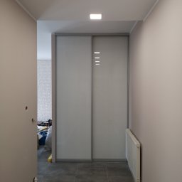 KEM Marcin Wielicki - Korytarz z szarą zabudową na wymiar z przesuwnymi drzwiami, widoczne trzy poziome kreski dekoracyjne, szare płytki podłogowe i grzejnik.