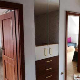 KEM Marcin Wielicki - Wysoka szafa z lustrzanymi drzwiami, białymi i brązowymi frontami szuflad, ze złotymi uchwytami, widziana w korytarzu obok otwartych drzwi do pokoju.