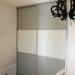 KEM Marcin Wielicki - Czteropanelowa szafa przesuwna z szarymi i białymi panelami, oświetlona przez czarny żyrandol z kryształkami, w pokoju z drewnianą podłogą i dekoracyjnymi gwiazdkami na suficie.