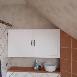 KEM Marcin Wielicki - Biała szafka łazienkowa z dwoma drzwiczkami i metalowymi uchwytami, zamontowana na ścianie z brązowymi płytkami w skośnej łazience na poddaszu. Na półce pod szafką stoją kosmetyki i miska.