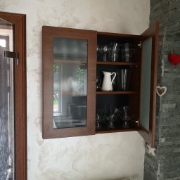 KEM Marcin Wielicki - Wisząca szafka z ciemnego drewna z przeszklonym frontem, wypełniona szkłem i dzbankami, zamontowana na ścianie z dekoracyjną fakturą, obok ściany z kamienia.