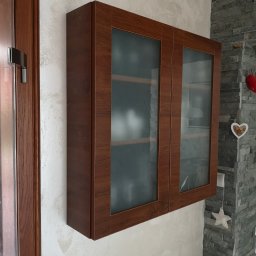 KEM Marcin Wielicki - Brązowa szafka wisząca z przeszklonymi drzwiczkami, zamontowana na ścianie o nierównej fakturze obok drewnianych drzwi i ściany z szarego kamienia dekoracyjnego.